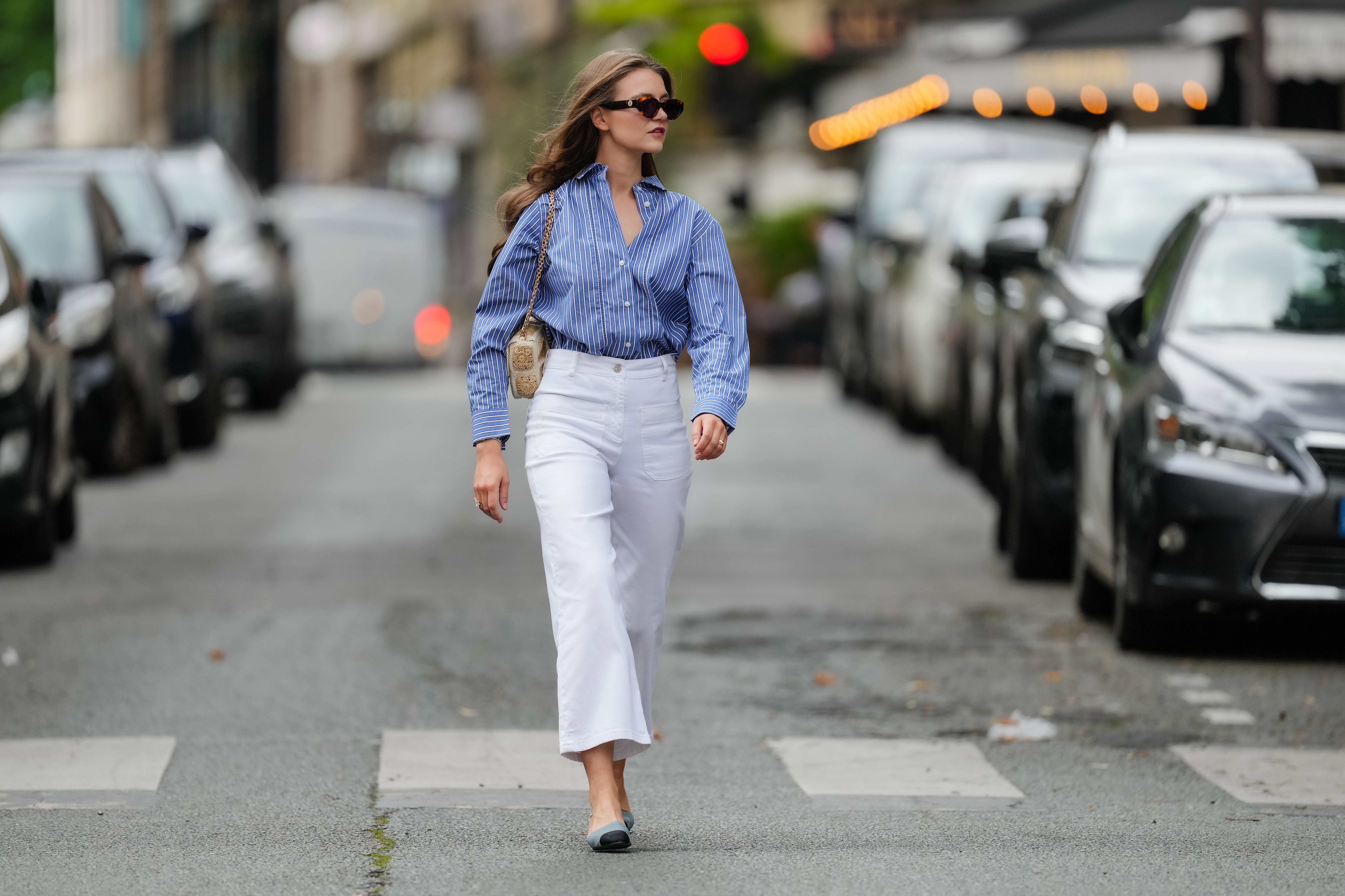 Segolene Hyppolite wears sunglasses, a blue and white oversized shirt from Tommy Hilfiger, a raffia beige bag from Vanessa Bruno, high waist white denim pants from Vanessa Bruno, Chanel slingback shoes in blue denim with black tips, during a street style fashion photo session, on May 20, 2024 in Paris, France.
