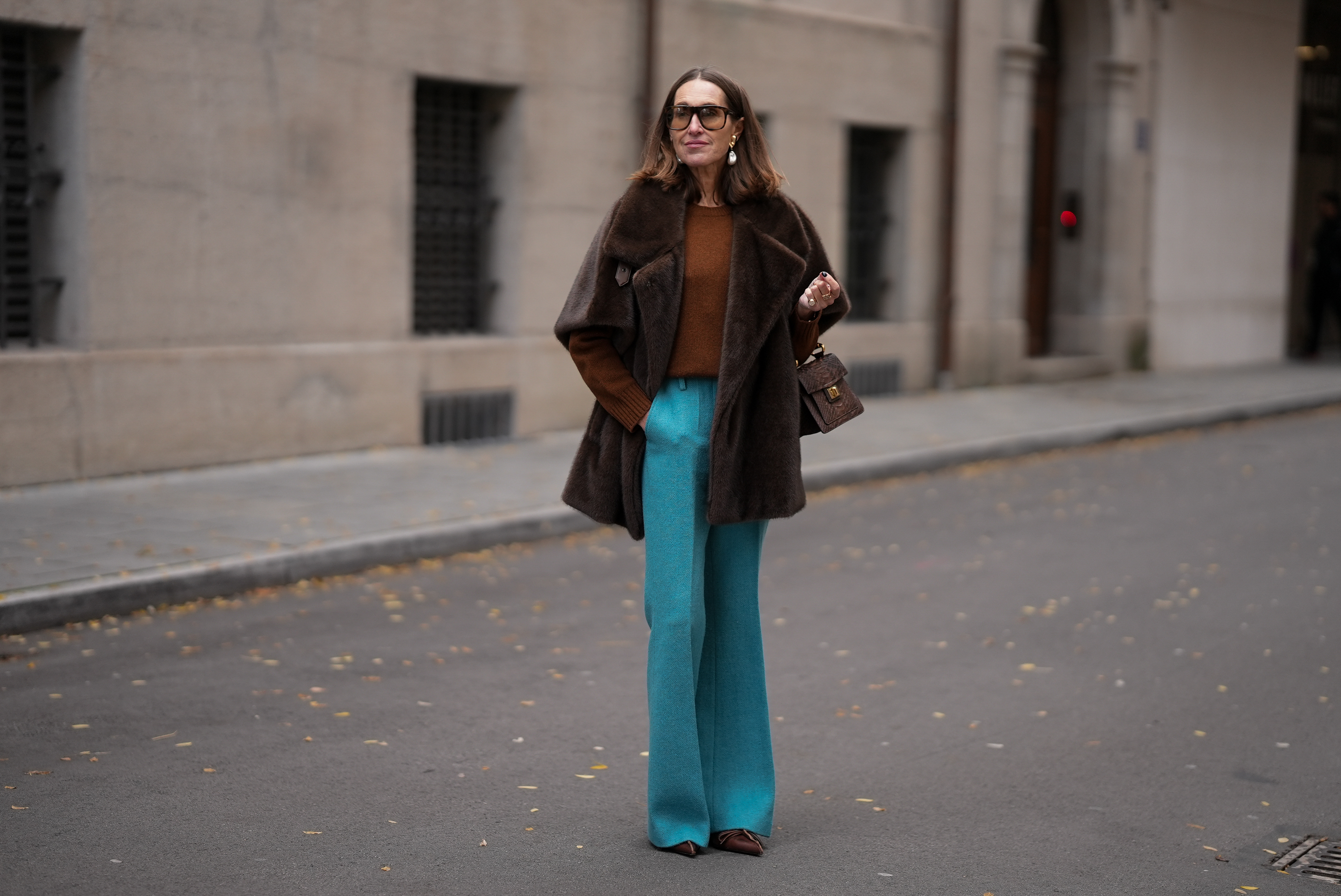 MUNICH, GERMANY - OCTOBER 28: Annette Weber is seen wearing a dark brown faux mink fur jacket with a plush, short sleeves, wide draped lapels and a relaxed cape like silhouette from Marina Rinaldi; a warm brown knit sweater with ribbed cuffs and crewneck from Prada; bright turquoise wool flare trousers with a soft structured boot cut and pressed front seams from Gembalies; dark brown leather pumps with a pointed toe, slim leather bow detail and a mid stiletto heel from Prada; a small structured top-handle bag in brown python leather with a hand-painted enamelled brass handle called ‘Greyhound Art Bag’ from Biagini 1988; oversized square sunglasses with a tortoiseshell patterned frame and warm brown gradient lenses from Saint Laurent; sculptural gold colored drop earrings with hanging pearls from Manuela Fumagalli; her brown hair is worn straight with a center part on October 28, 2025 in Munich, Germany. (Photo by Moritz Scholz/Getty Images)