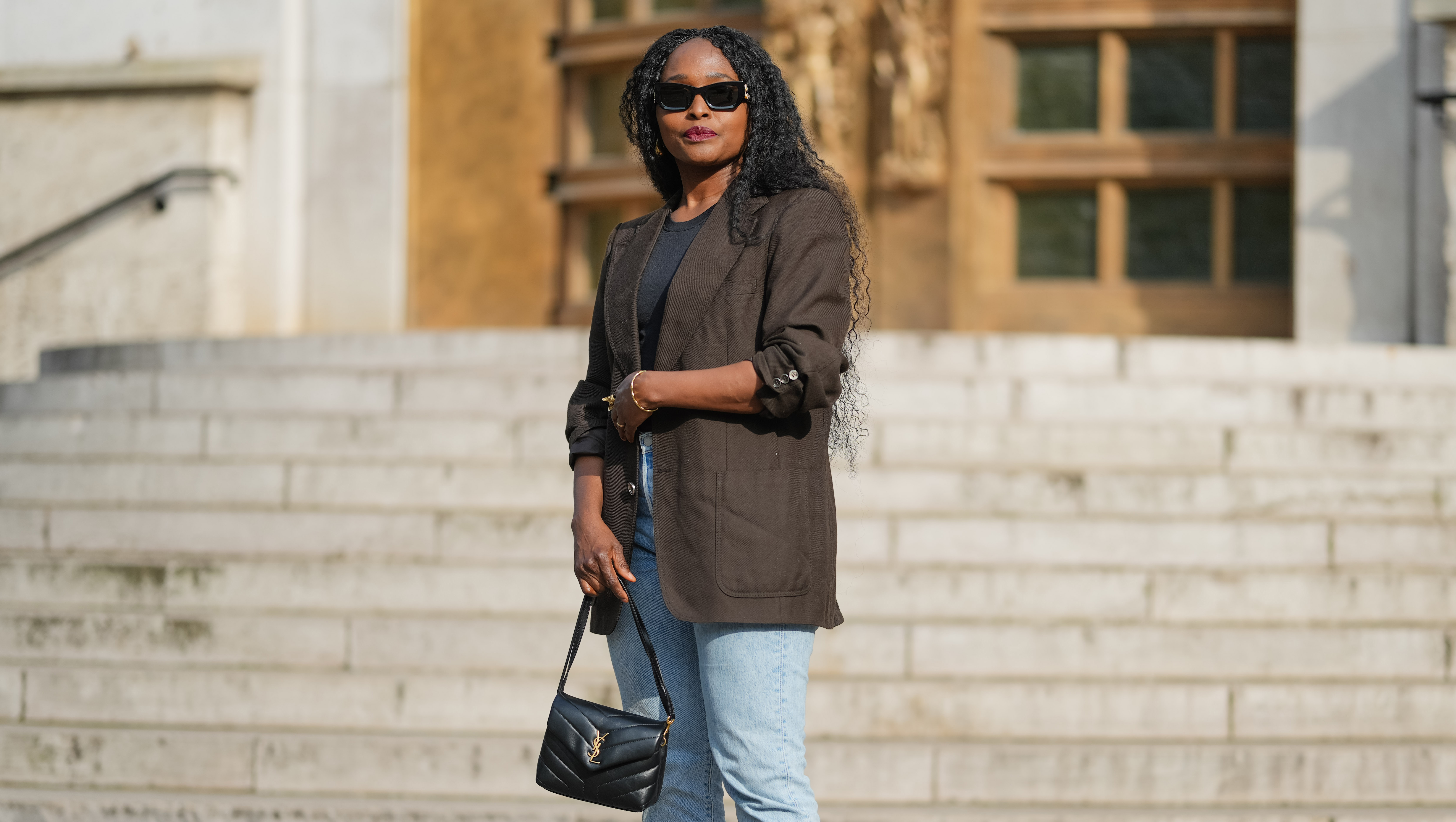 PARIS, FRANCE - MAY 02: Carrole Sagba wears long, curly black hair parted in the middle. Black sunglasses cover the eyes, and red lipstick is applied. A dark brown blazer jacket made of a textured material is worn over a black top. Light blue denim jeans with a straight cut are paired with black studded slip-on shoes. A black leather YSL Saint Laurent handbag with a visible gold logo is carried. The image is a full body shot, during a street style fashion photo session, on May 02, 2025 in Paris, France. (Photo by Edward Berthelot/Getty Images)