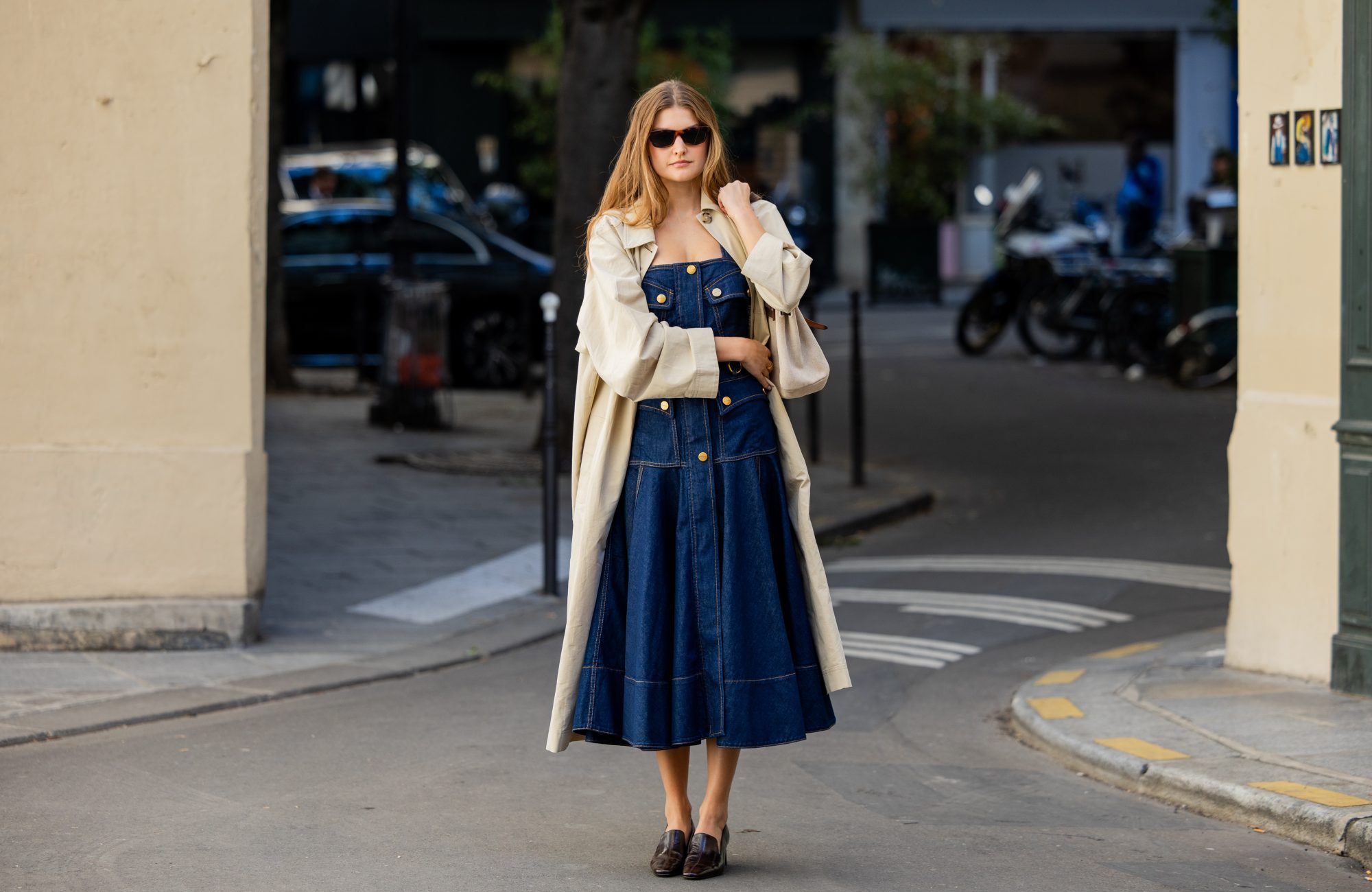 PARIS, FRANCE - OCTOBER 05: Sara Walker wears a dark wash denim midi dress with brass button details, a light beige trench coat, dark brown sunglasses and black patent leather block heel loafers outside Zimmermann on October 05, 2026 in Paris, France. (Photo by Christian Vierig/Getty Images)