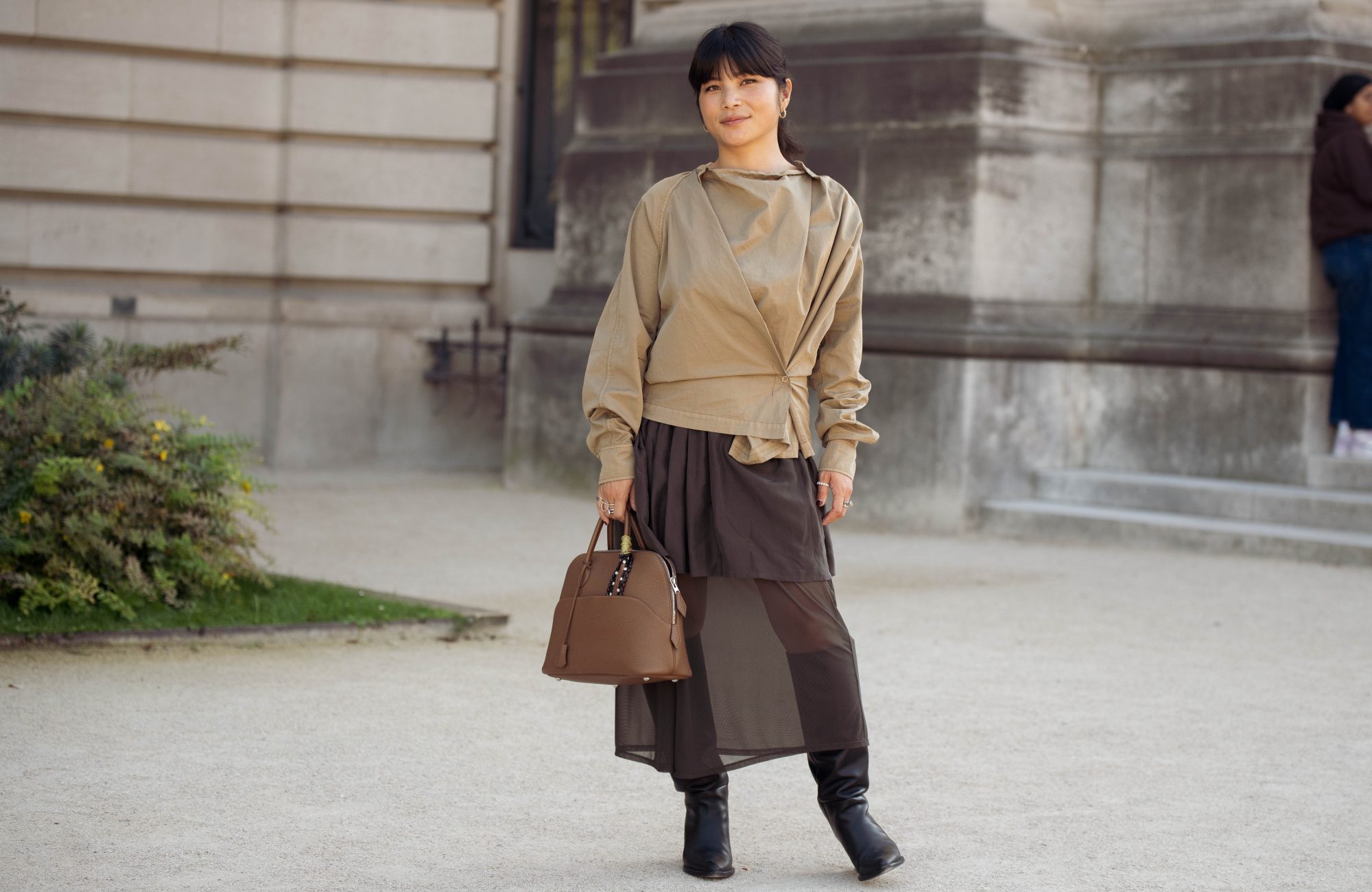 PARIS, FRANCE - OCTOBER 05: Elie Inoue wears a beige draped wrap-style top with a layered dark brown skirt featuring sheer panels, carries a structured brown top-handle bag, and black knee-high boots outside Gabriela Hearst show during Day Eight of Paris Fashion Week - Womenswear Spring/Summer 2027 on October 05, 2026 in Paris, France. (Photo by Raimonda Kulikauskiene/Getty Images)